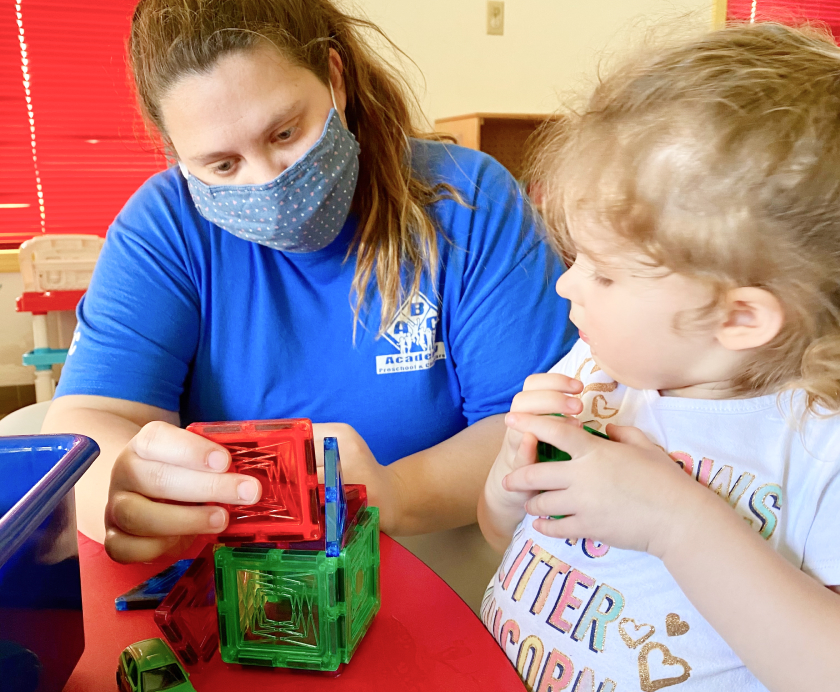 A young woman wearing a mask assisting a little girl with blocks.