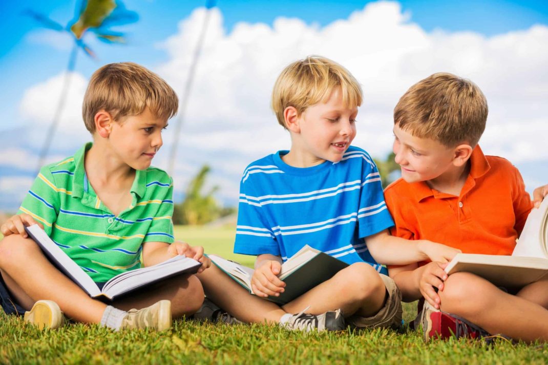 Three boys sitting on the grass reading books.