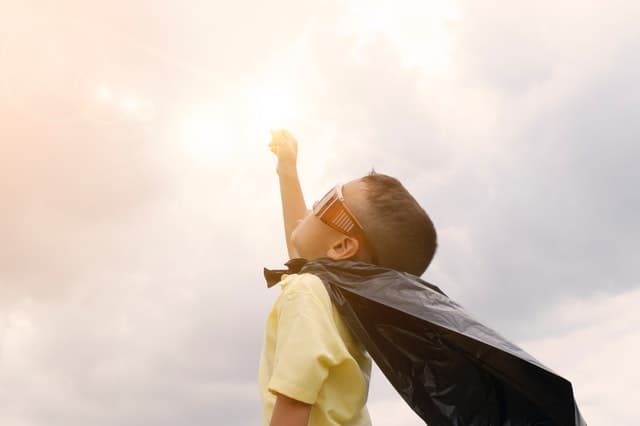 A young boy in a superhero cape is reaching for the sun.