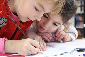 Two girls are sitting at a table and writing in a book.