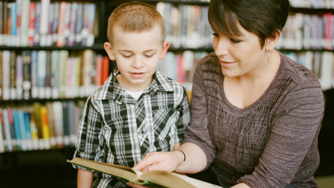 A teacher guiding a little boy with a chapter book.