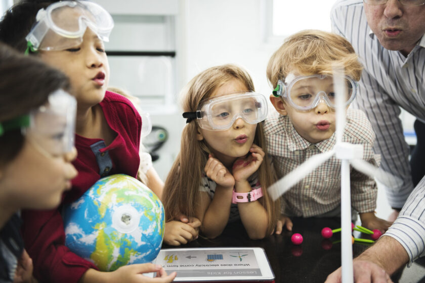 Children wearing safety goggles observe a wind turbine model in a classroom. One child holds a globe, and an adult is partially visible guiding them. Educational materials are on the table.