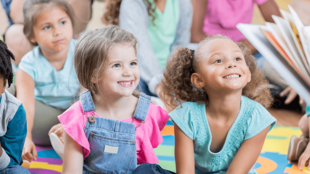 A group of young children sits on the floor in a classroom, attentively listening to someone reading a book. Two girls in the front are smiling.