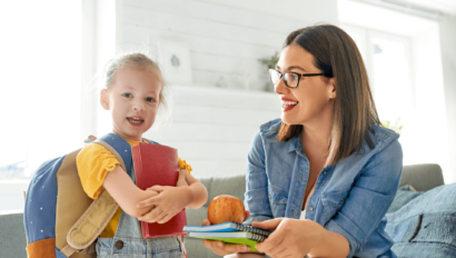A child with a backpack holds books while a woman in glasses smiles, offering fruit and more books, in a bright room.