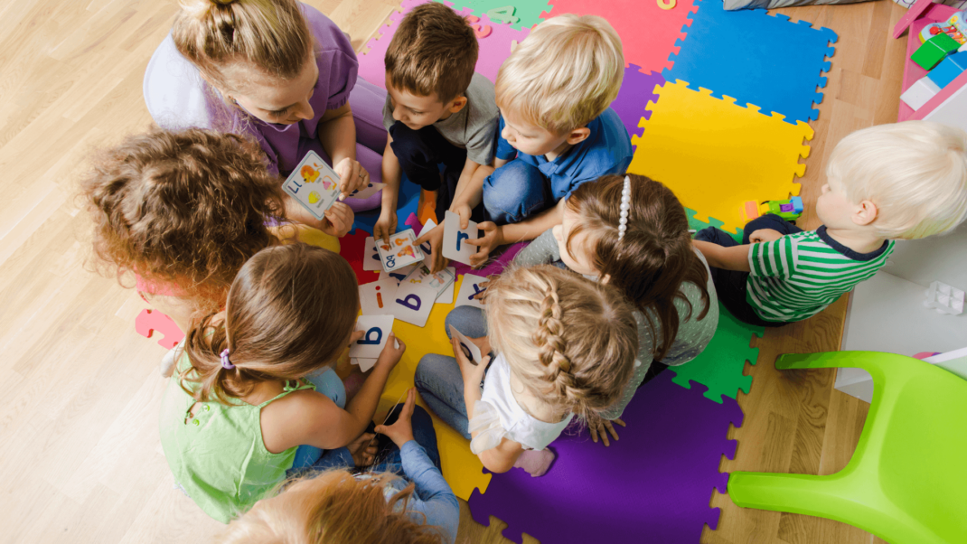 Children sitting on colorful foam mats engage in a group learning activity with letter cards, guided by an adult.