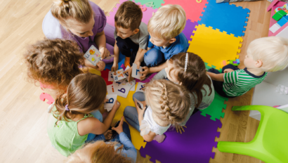 Children sitting on colorful foam mats engage in a group learning activity with letter cards, guided by an adult.