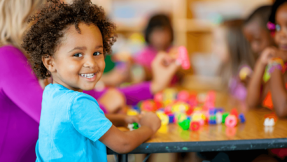 Child in a blue shirt smiles at the camera while sitting at a table with other children, playing with colorful toys and blocks in a classroom setting.