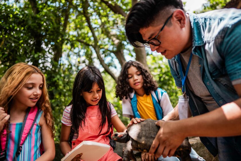 A man shows a turtle to three children outdoors; one child holds a notebook, and the group observes the turtle closely in a wooded area.