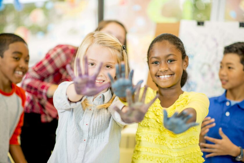 Four children smile and show their paint-covered hands toward the camera in a bright classroom setting.