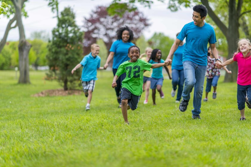 A group of children and adults are running together on grass in a park, appearing active and cheerful.